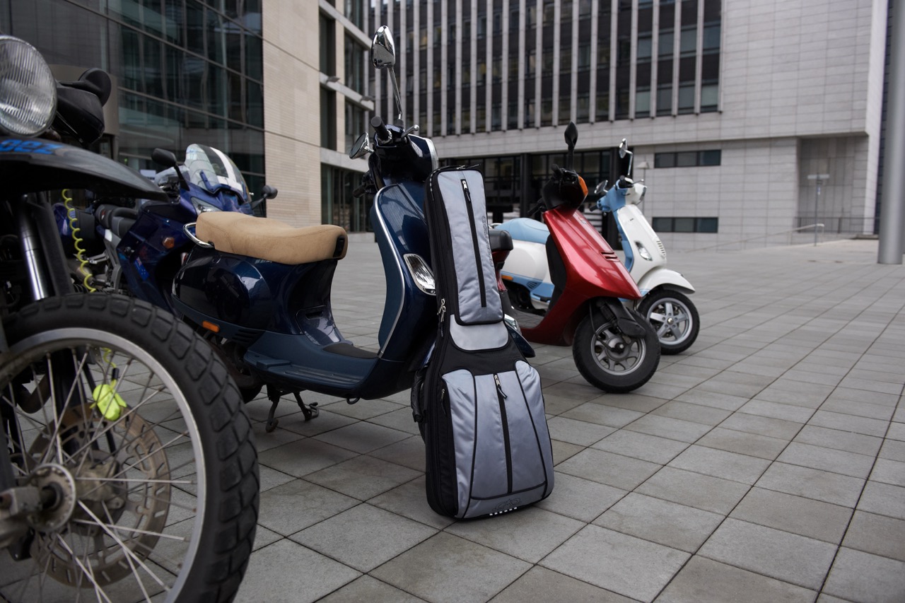 Gray and black MADAROZZO guitar gig bag standing next to parked scooters and motorcycles in a paved urban courtyard.