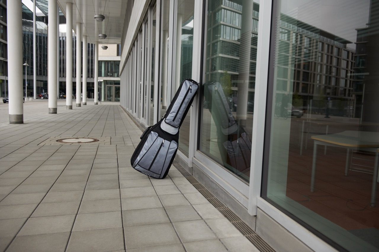 Gray and black MADAROZZO guitar gig bag leaning against the glass facade of a contemporary building along a covered colonnade.