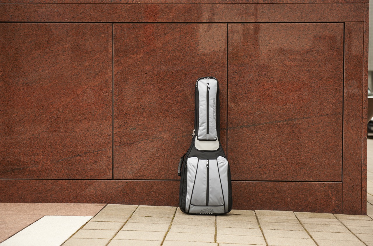 Gray and black MADAROZZO guitar gig bag standing in front of a large reddish-brown stone wall on tiled pavement.