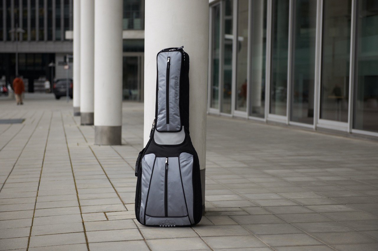 Gray and black MADAROZZO guitar gig bag leaning against a white pillar in a modern urban plaza with glass buildings.