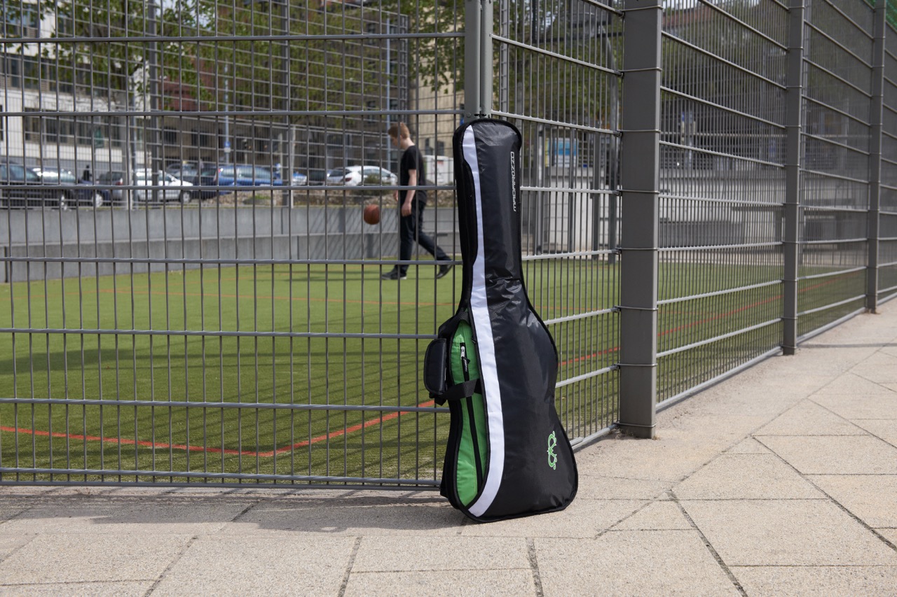 Black MADAROZZO guitar gig bag with green accents and white stripe leaning against a metal fence beside a green sports field.