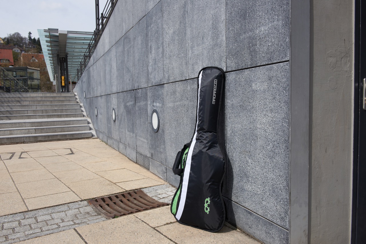 Black and green MADAROZZO guitar gig bag leaning against a textured gray concrete wall near outdoor stairs.