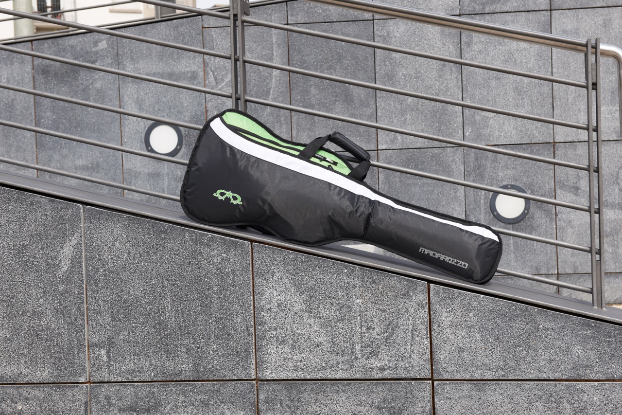 Black and green MADAROZZO guitar gig bag lying horizontally on a metal railing against a tiled wall.