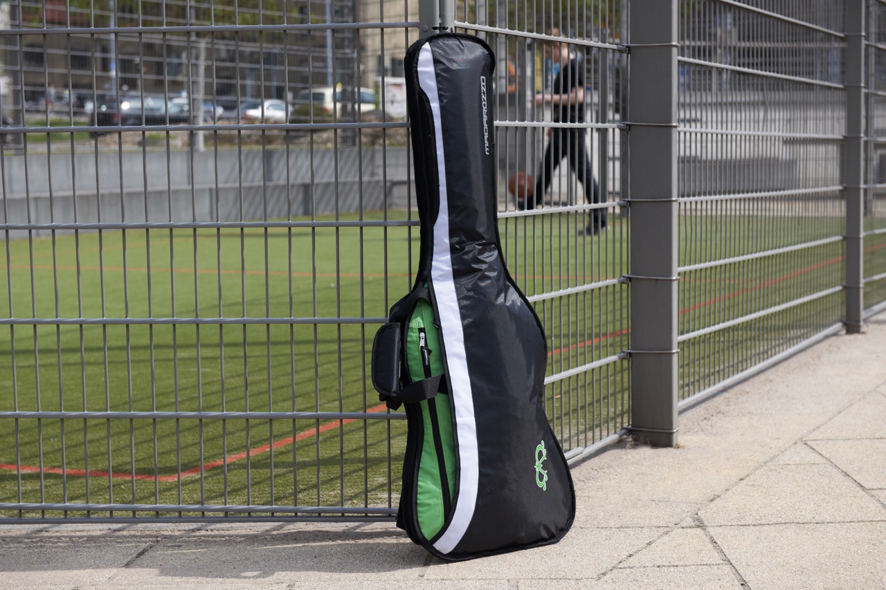 Black MADAROZZO guitar gig bag with green accents leaning against a metal fence with an artificial turf sports field behind it.